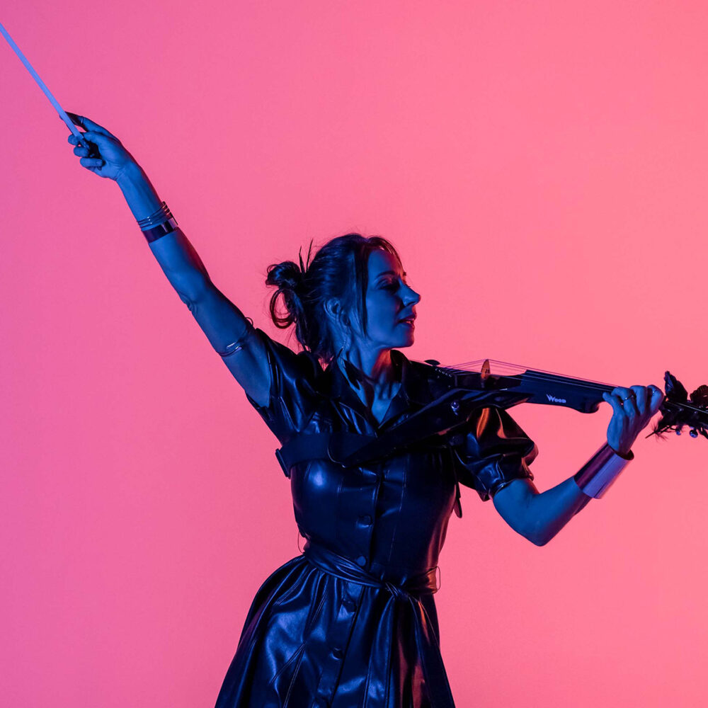 A woman in a black dress plays an electric violin, holding the bow high with one arm. She is illuminated by dramatic blue and pink lighting against a vibrant pink background.