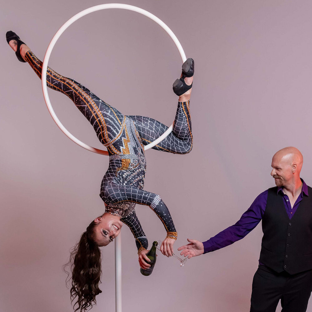 A woman in a patterned bodysuit hangs upside down from a white aerial hoop, smiling, while a bald man in a purple shirt and black vest stands beside her with an outstretched hand.