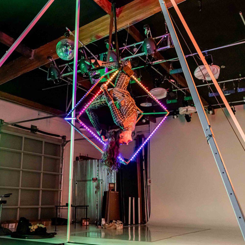 An aerial artist in a sparkling outfit hangs upside down inside a glowing, neon-lit cube suspended from the ceiling in a studio with stage lights and exposed beams.