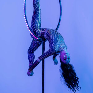 An aerial artist in a sparkling bodysuit performs an aerial hoop act, hanging upside down with one leg hooked over the hoop and her hair flowing downward, illuminated by vibrant blue and purple lights.