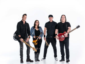 Four musicians stand against a white background. Two men hold electric guitars, a woman holds a violin, and another man holds drumsticks. They are dressed in black and appear to be part of a rock band.