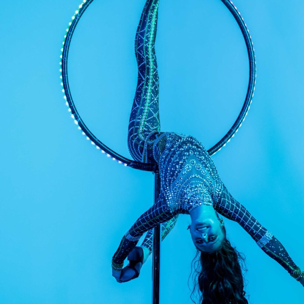 An aerial artist in a sparkling bodysuit hangs upside down from an aerial hoop against a blue background, with one leg hooked over the hoop and hair cascading downward.