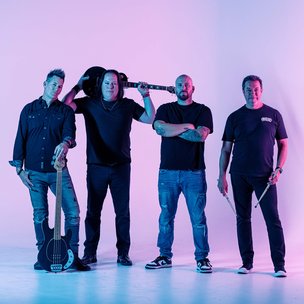 Four men stand together in a studio with purple and blue lighting. Two hold guitars, one rests a guitar on his shoulder, and one holds drumsticks. They wear casual clothing and pose confidently.