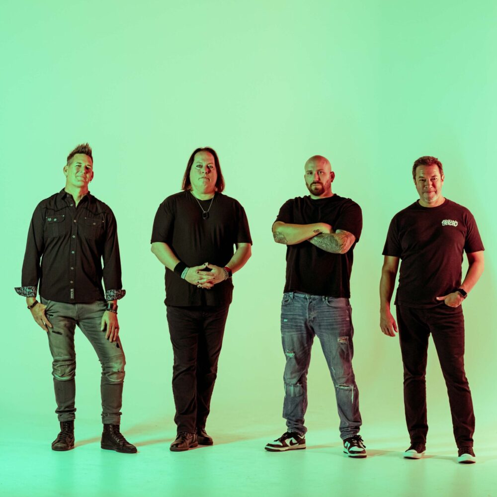 Four men stand side by side in a brightly lit studio with a green gradient background. They wear casual clothing, including dark shirts and jeans, and pose facing the camera.