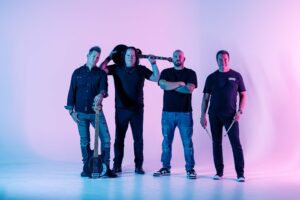 Four men stand side by side in a brightly lit studio with pink and blue tones. Two hold guitars, one holds drumsticks, and one has a guitar resting on his shoulder. They wear casual, dark clothing and look toward the camera.