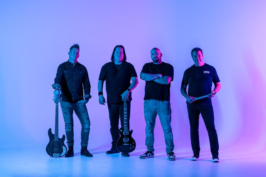 Four men stand in a row against a blue and pink gradient background. Two hold guitars, one stands with arms crossed, and one holds drumsticks. They all wear dark clothing and look at the camera.