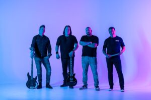 Four men stand in a row against a blue and pink gradient background. Two hold guitars, one stands with arms crossed, and one holds drumsticks. They all wear dark clothing and look at the camera.