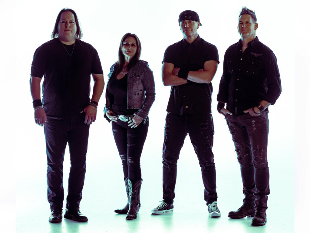 Four people stand side by side facing the camera, dressed in dark, casual clothing. One woman is second from the left, and the others are men. The background is bright white, creating a silhouette effect.