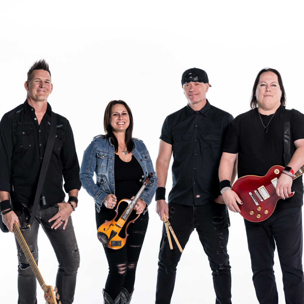 Four musicians stand together against a white background, holding instruments including guitars, drumsticks, and a violin. They are dressed mostly in black, smiling and facing the camera.