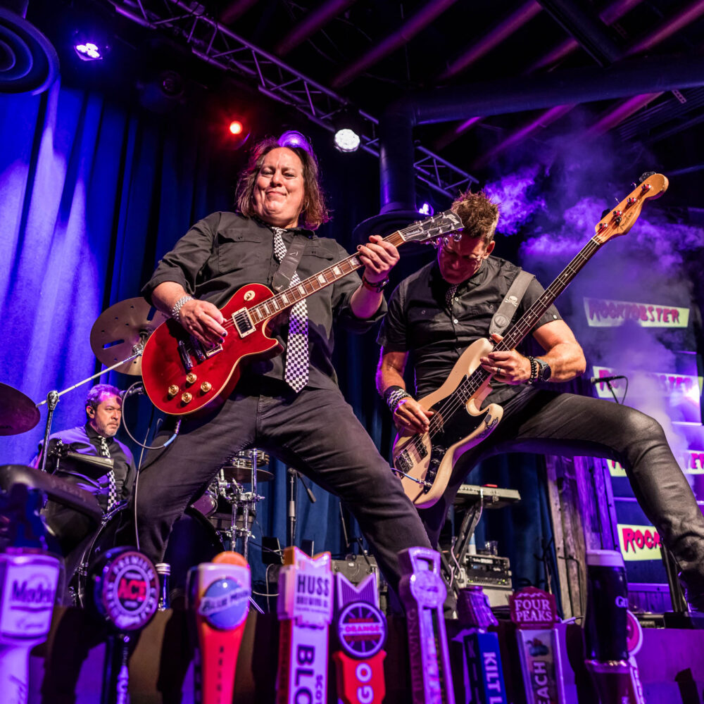 A guitarist and bassist on stage rocking live, with the drummer in the far background