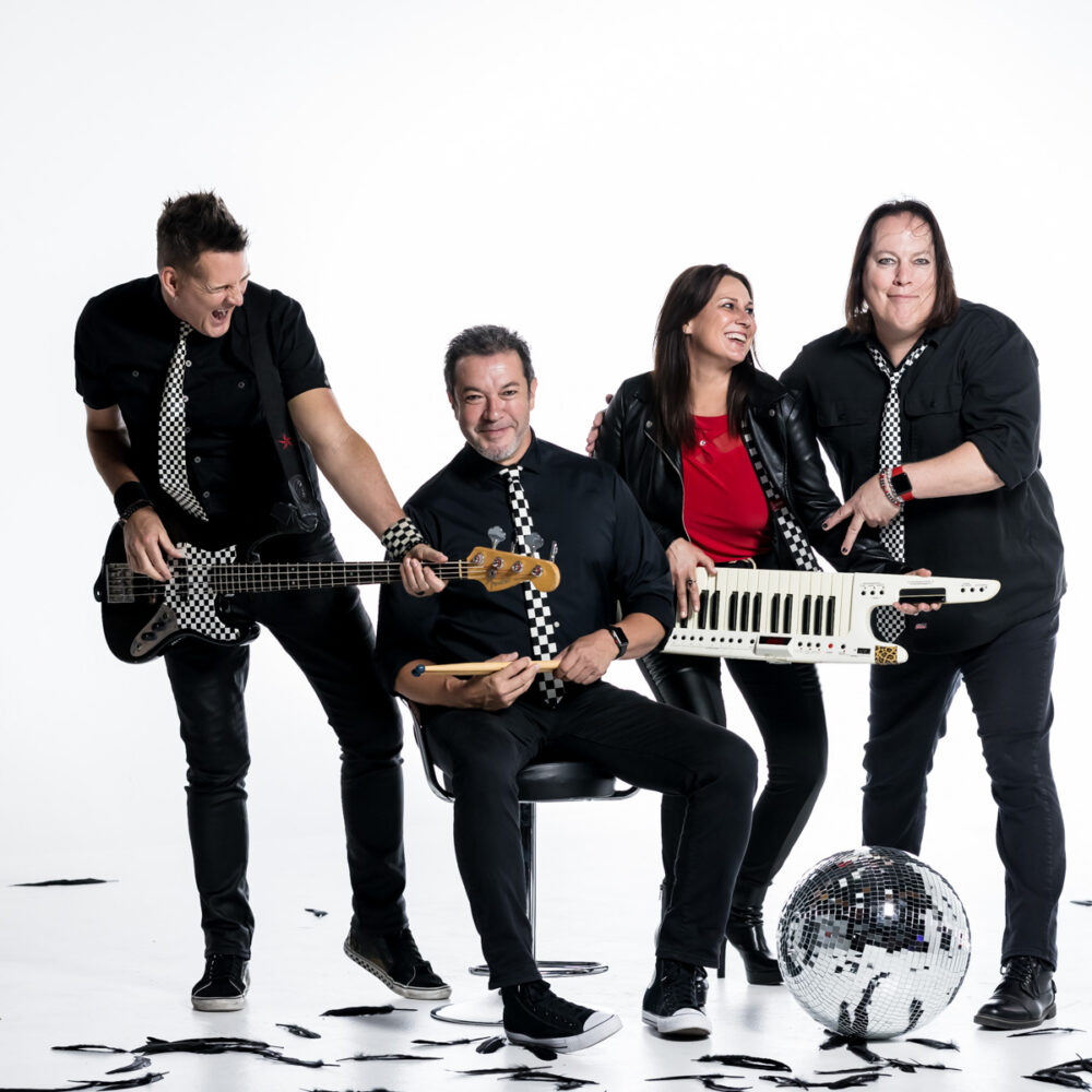 Four people in black outfits and checkered ties pose playfully with musical instruments—a bass, a keytar, and drumsticks—against a white background, with a disco ball and black confetti on the floor.