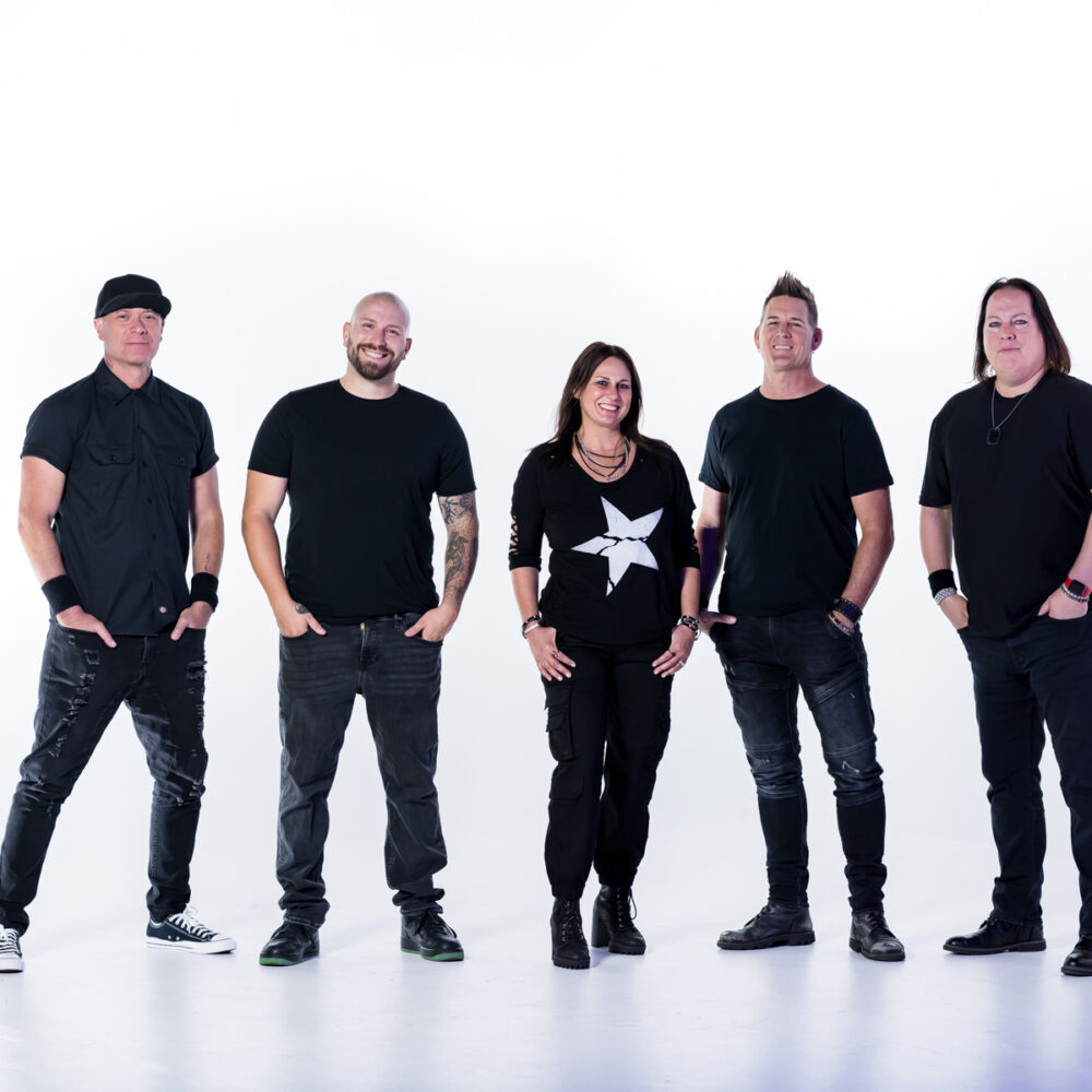 Five people stand in a line against a white background, all wearing black outfits and smiling at the camera. The person in the center wears a shirt with a large white star.